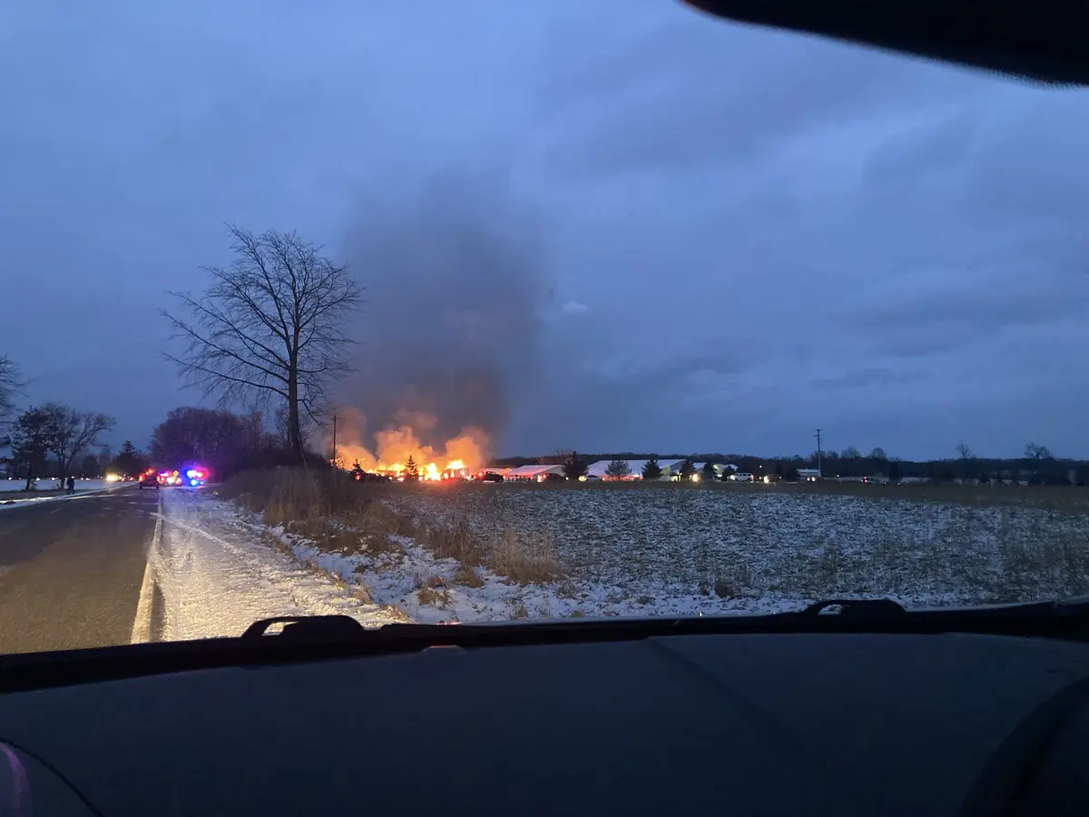 A photo taken from inside a car shows a large fire burning in a snowy field at dusk, with emergency vehicles and flashing…