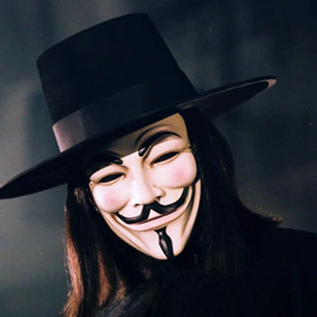 A person with long dark hair wears a black top hat and a white Guy Fawkes mask with a wide grin and mustache, set against a…