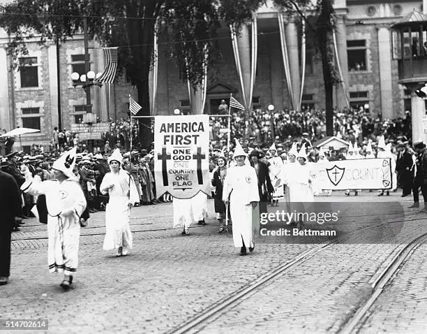 A black-and-white historical photo shows members of the Ku Klux Klan marching in a parade, wearing white robes and hoods…