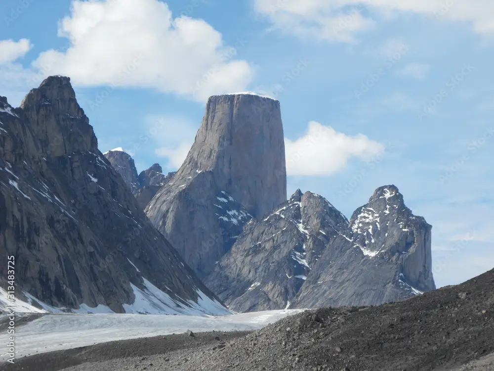 A scenic mountain landscape with a prominent, steep, gray rock formation rising above snow-dusted peaks and a glacier in the…
