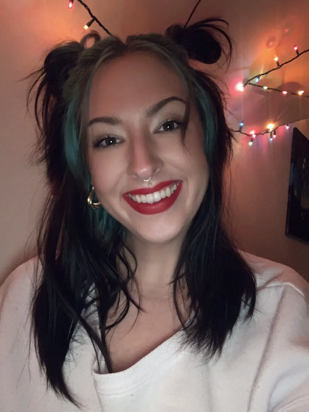 A smiling young woman with dark hair featuring teal streaks, styled in two messy buns, wearing red lipstick, a nose ring…