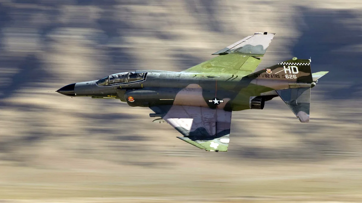 A military jet, likely a USAF F-4 Phantom II, is captured in high-speed flight with a blurred background indicating rapid…