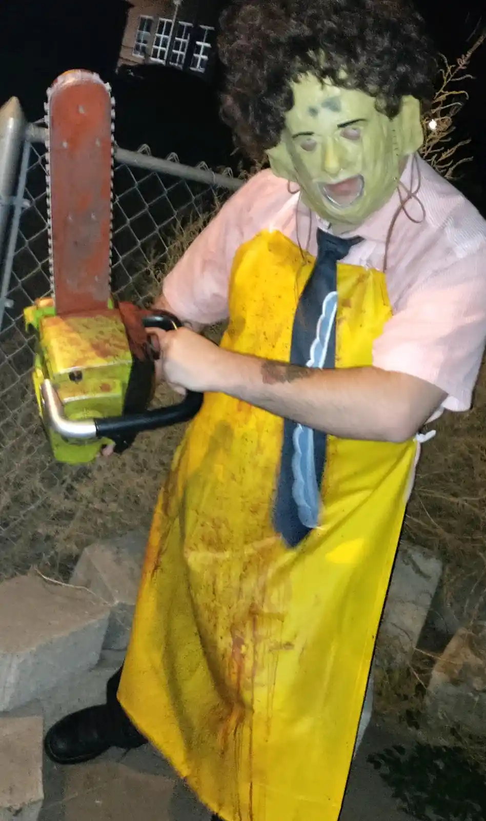 A person wearing a green mask, curly wig, pink shirt, and yellow apron covered in fake blood holds a chainsaw. The chainsaw…