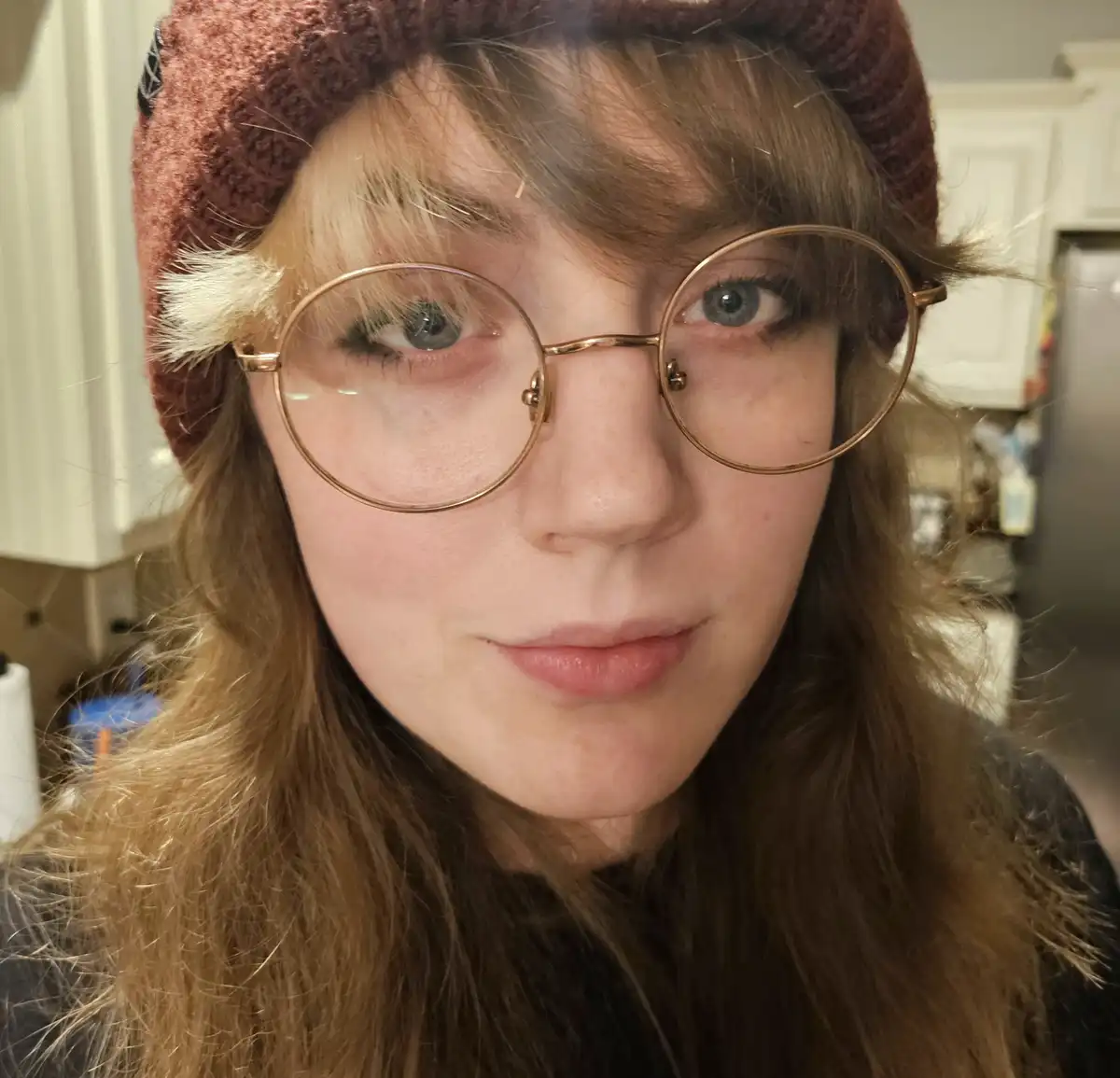 A close-up selfie of a person with long brown hair, wearing a maroon knit beanie with a white fluffy pom-pom on the side and…