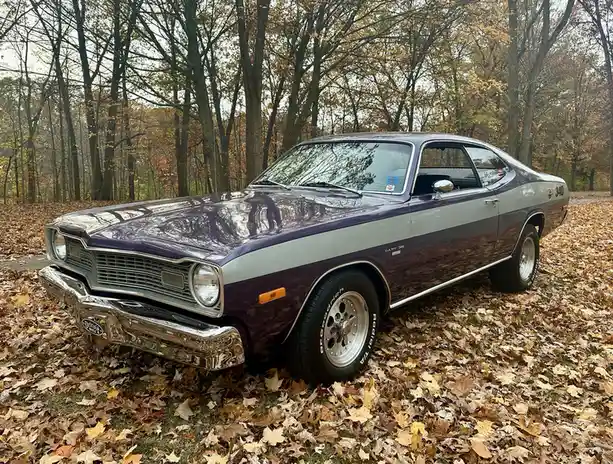 A shiny purple and silver 1970s Dodge Dart 340 muscle car is parked in a forest covered in fallen autumn leaves. The car has…