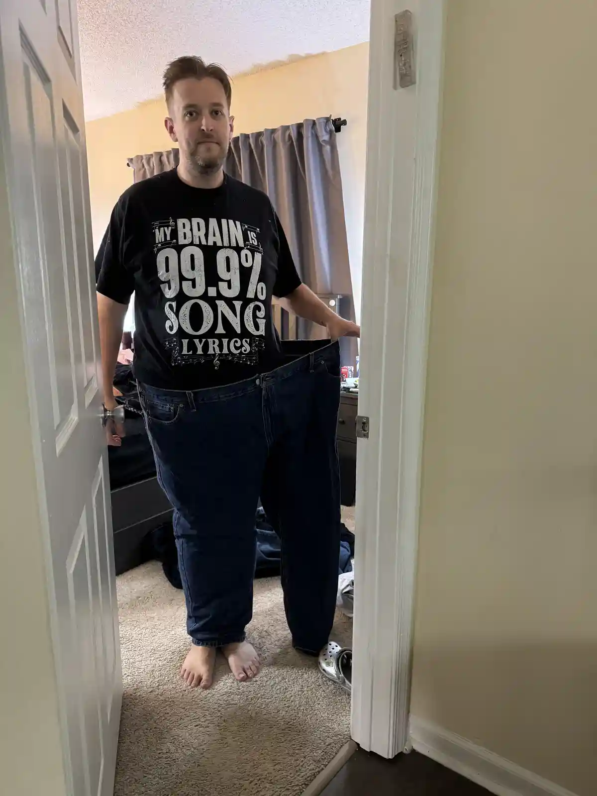 A man stands in a doorway wearing a black t-shirt that reads 'MY BRAIN IS 99.9% SONG LYRICS' surrounded by musical notes…