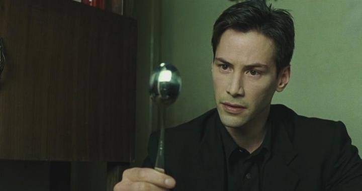 a man in a black shirt holds a metal spoon and stares intently at the viewer with a serious, slightly confused expression…