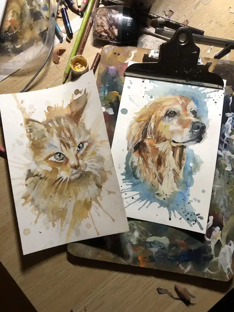 Two watercolor paintings of a cat and a dog are displayed on a cluttered artist's desk, surrounded by pencils, paintbrushes…