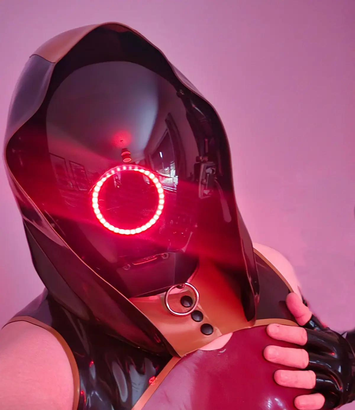 A person wearing a glossy black, futuristic helmet with a glowing red ring around the eye area, set against a pink-lit…