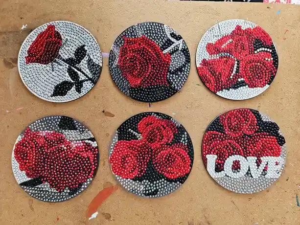 Six circular coasters made with rhinestones depict red roses and the word 'love' in white letters, arranged on a brown…