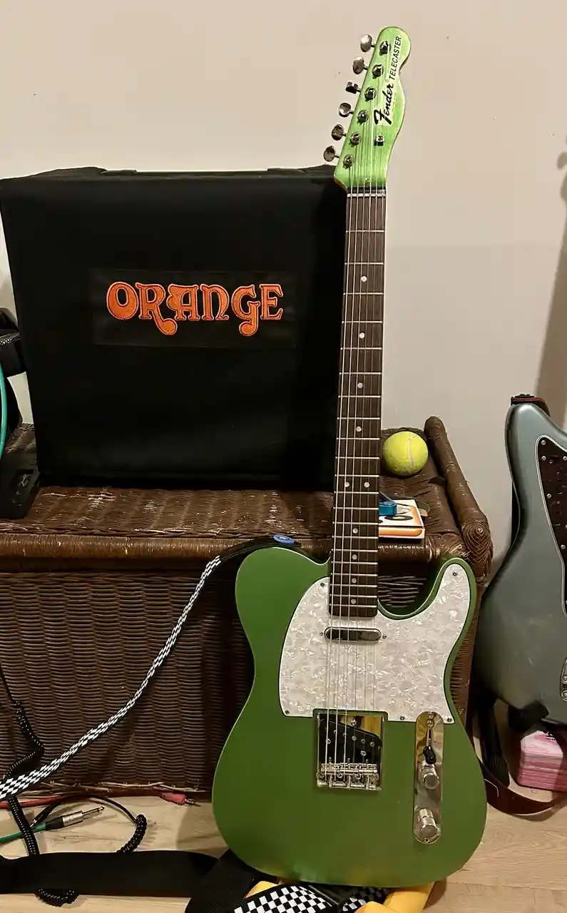 A green Fender Telecaster electric guitar leans against a black amplifier with 'ORANGE' embroidered in orange script on its…