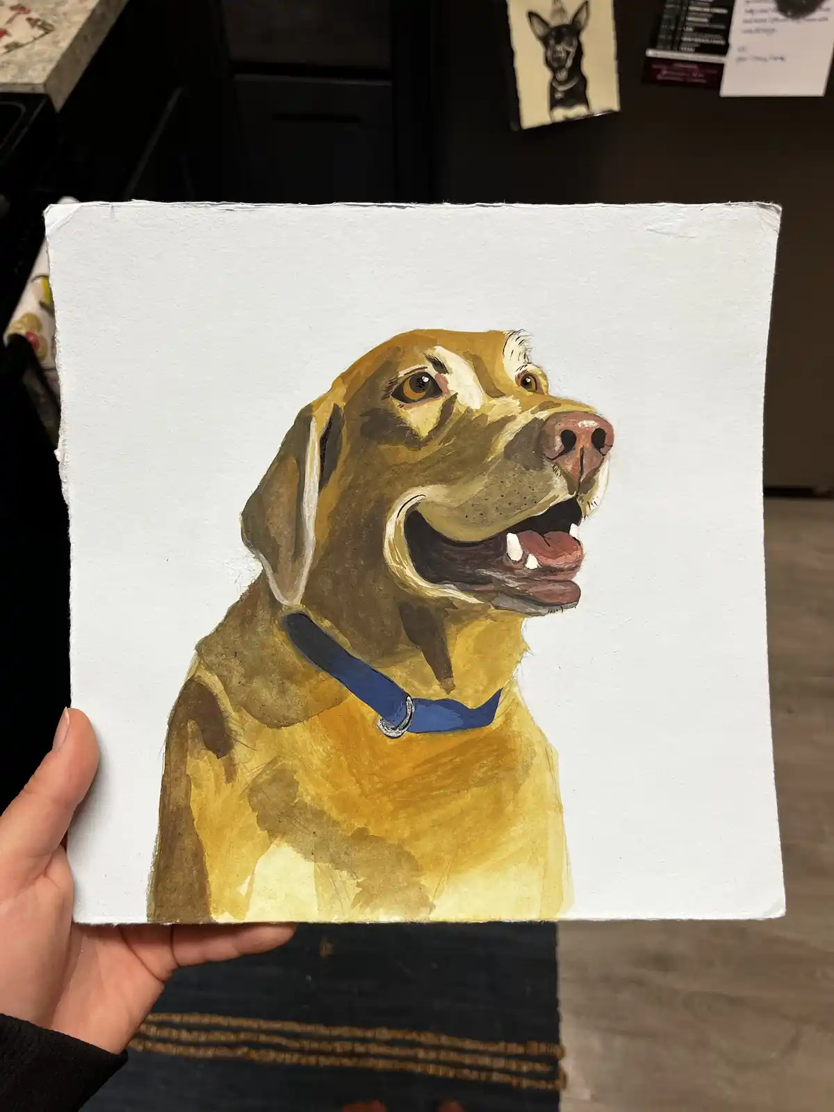 A hand holds up a watercolor painting of a yellow dog with a blue collar, looking upward with its mouth open as if smiling…
