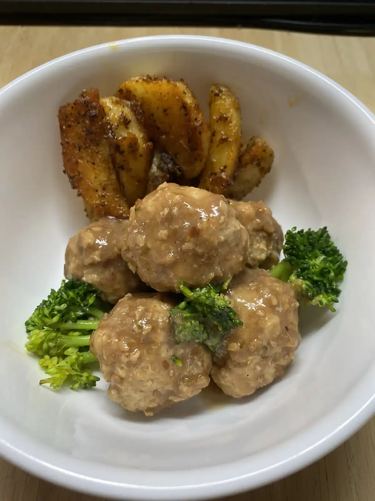 A white bowl contains several meatballs covered in a brown sauce, accompanied by roasted potato wedges and green broccoli…
