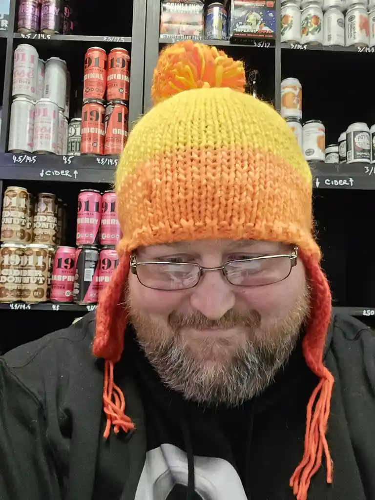 A man with glasses and a beard wears a bright yellow and orange knit hat with ear flaps and a pom-pom, smiling slightly…