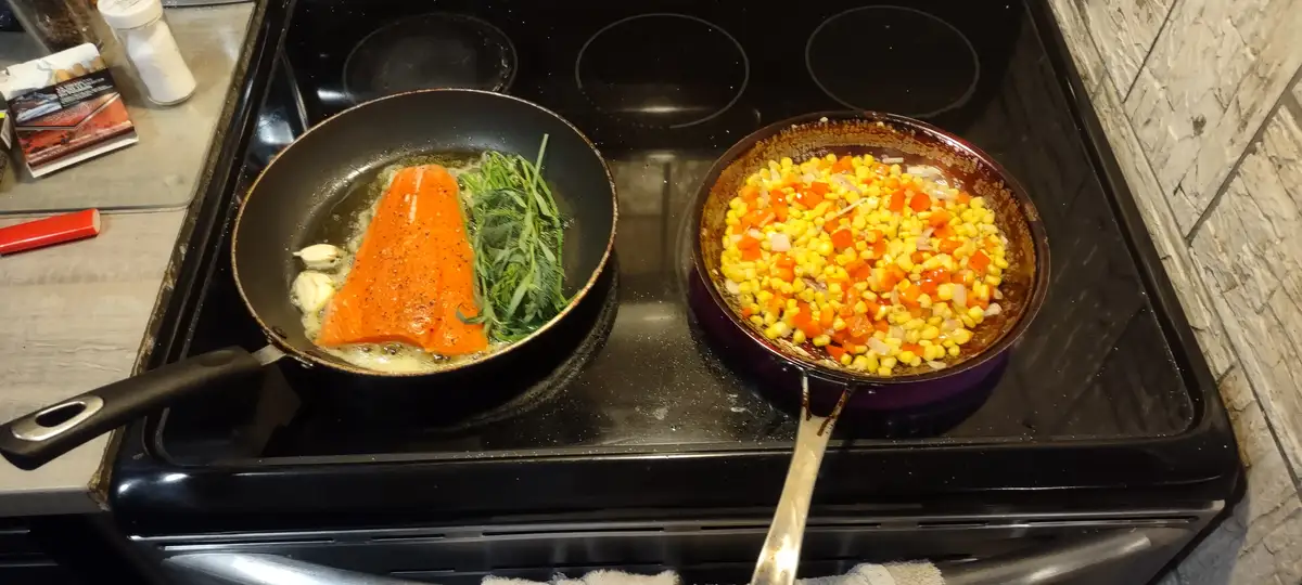 Two pans sit on a stovetop: one with a salmon fillet cooking in butter with garlic and herbs, the other with corn, red…