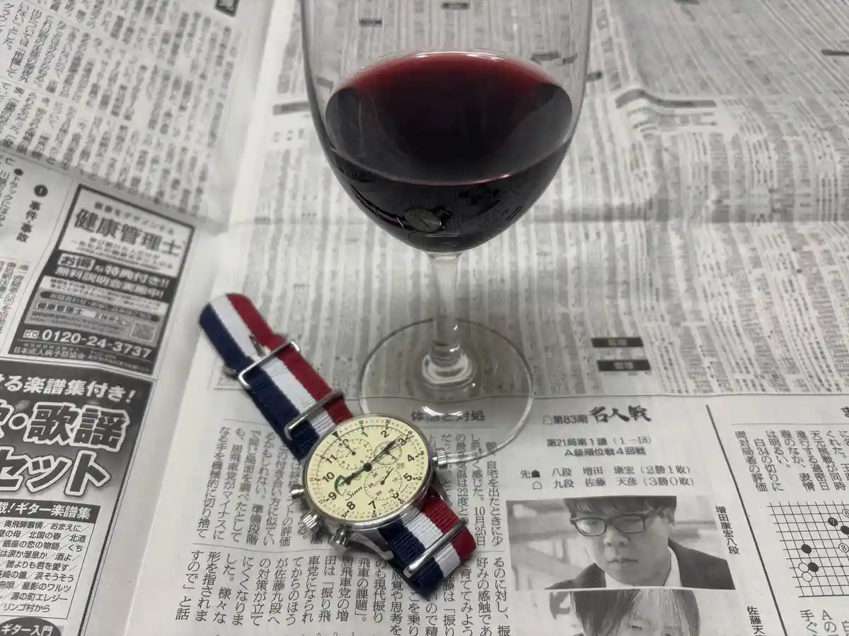 A glass of red wine and a watch with a red, white, and blue strap rest on a Japanese newspaper. The watch face is…