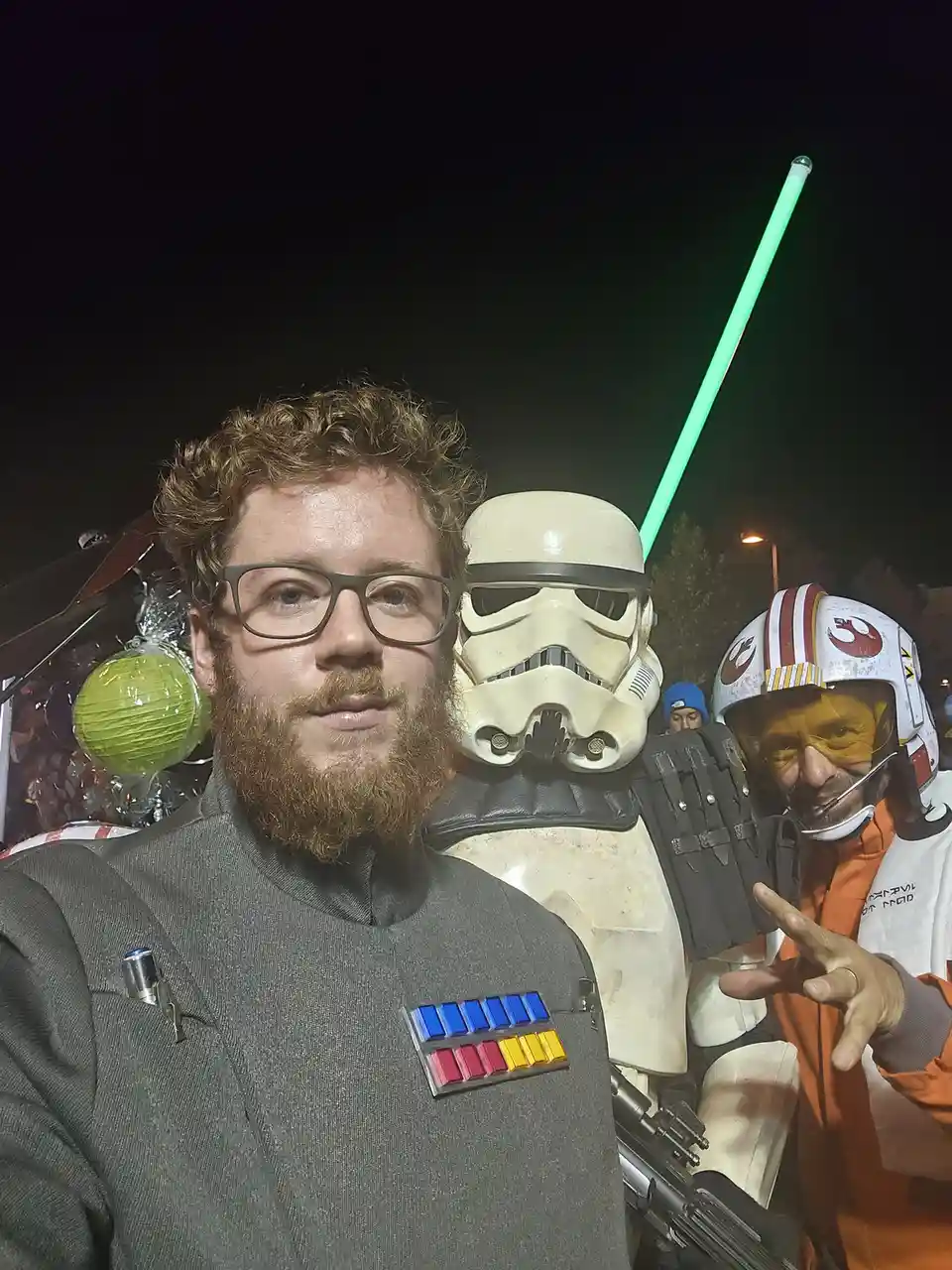 A man with glasses and a beard in a gray Star Wars uniform with colored rank insignia takes a selfie at night. Behind him…