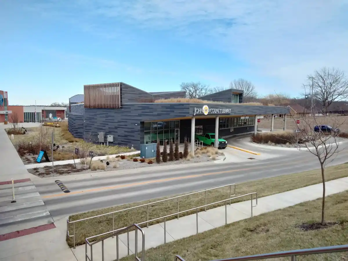 An exterior photo of the modern Johnson County Library building with dark gray siding, a green pickup truck parked under its…