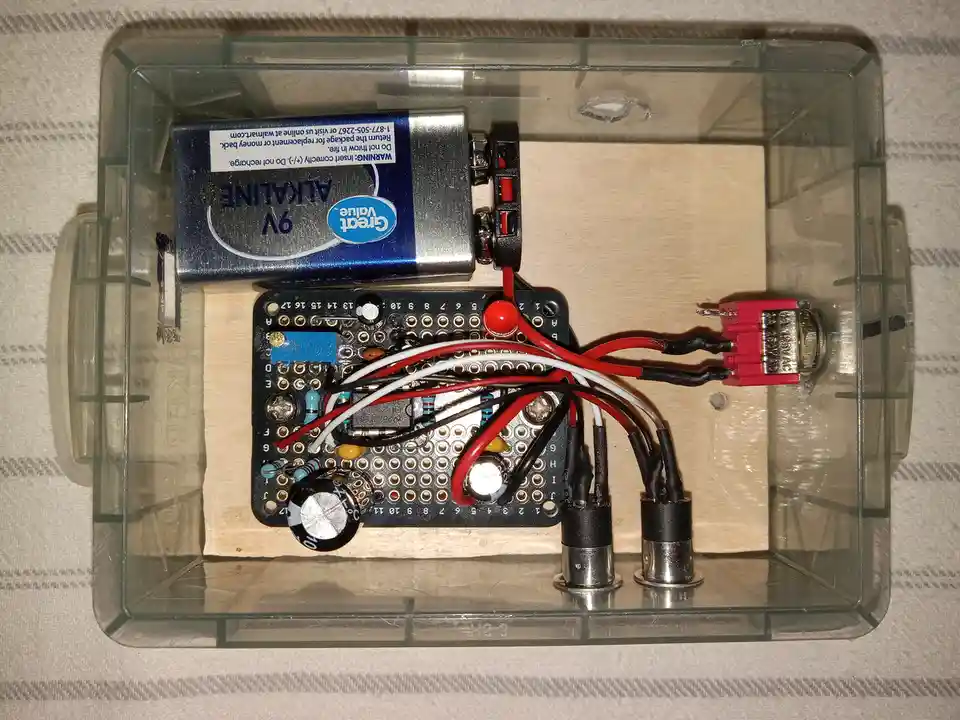 A transparent plastic box contains a 9V battery, a breadboard with various electronic components, wires, and a small…