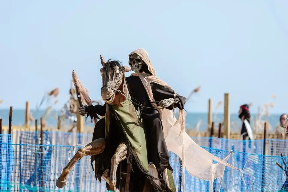 A person dressed as the grim reaper, complete with skull mask and hooded robe, is riding a skeletal horse on a beach. The…