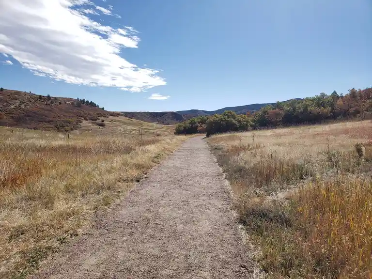 A wide gravel trail winds through a dry, grassy field under a blue sky with scattered clouds, flanked by autumn-colored…