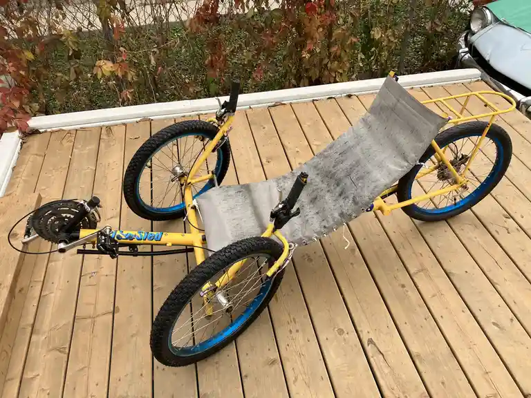 A yellow recumbent bicycle with a long gray fabric seat is lying on its side on a wooden deck. The bike has three wheels…