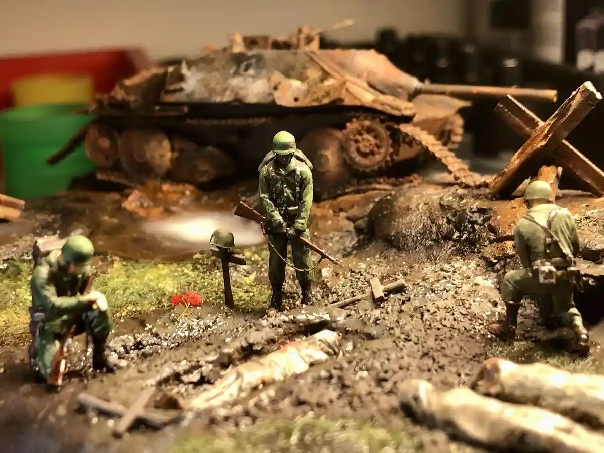 A diorama of miniature World War II soldiers in green uniforms amidst a battlefield with a destroyed tank, crosses, and…