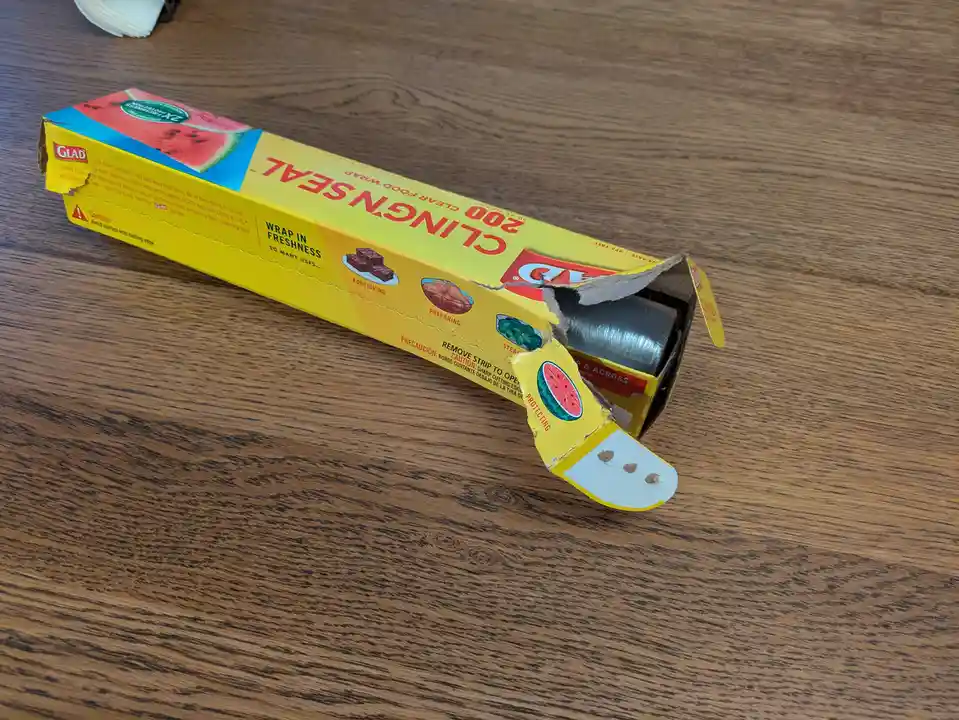 A yellow Glad Cling'n Seal food wrap box lies on a wooden table, torn open at the end to reveal the roll of plastic wrap…
