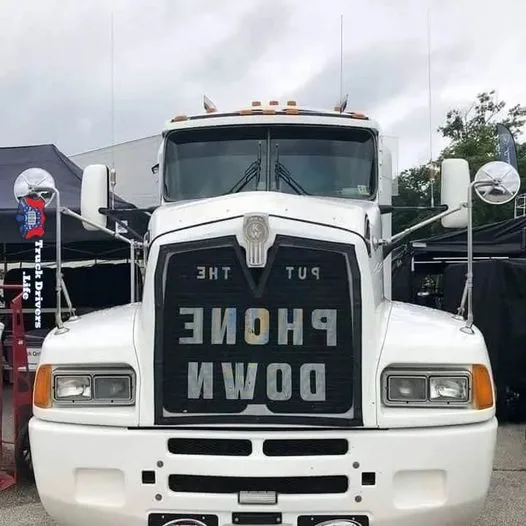 A white semi-truck's front grille has the words 'PUT THE PHONE DOWN' spelled out in large, reflective letters. The truck is…