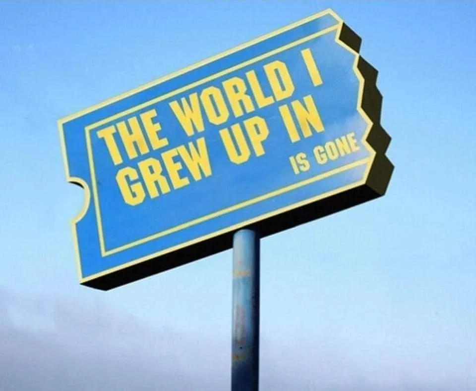 A blue, ticket-shaped sign with yellow text mounted on a pole against a clear sky. The text reads 'THE WORLD I GREW UP IN IS…