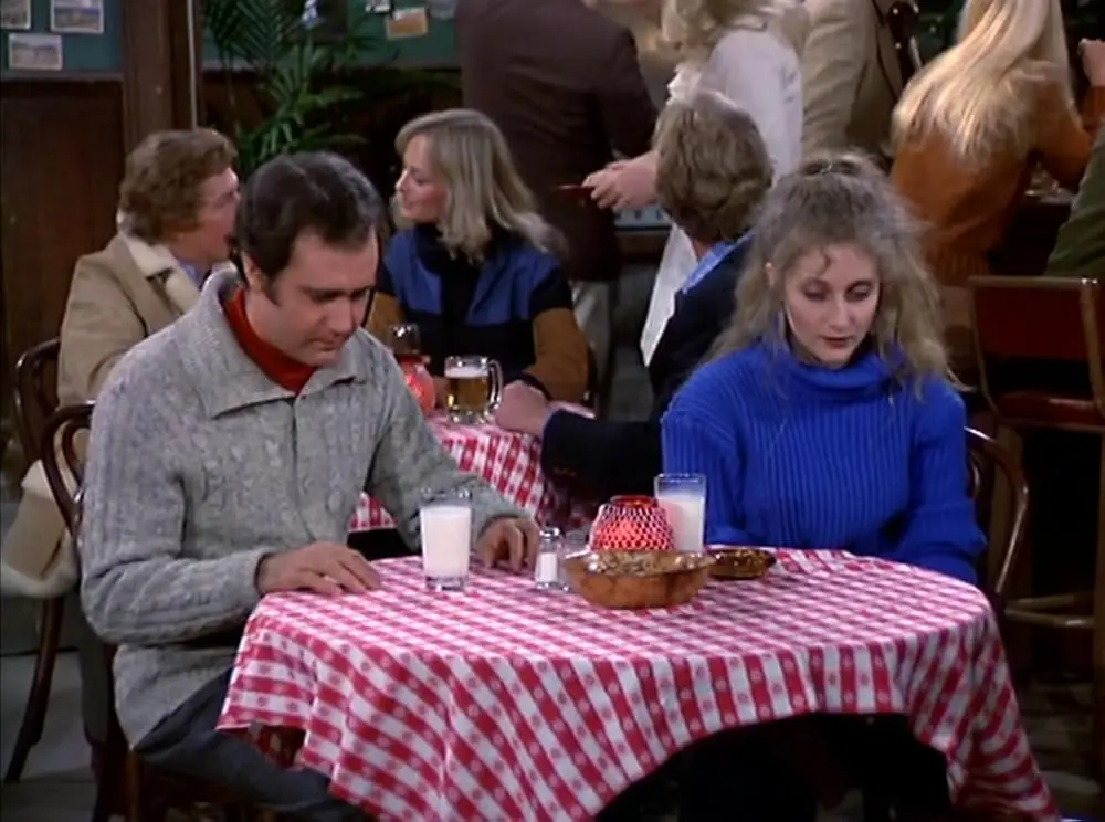 A man and woman sit at a red-and-white checkered table in a restaurant, both looking downcast and avoiding eye contact. The…