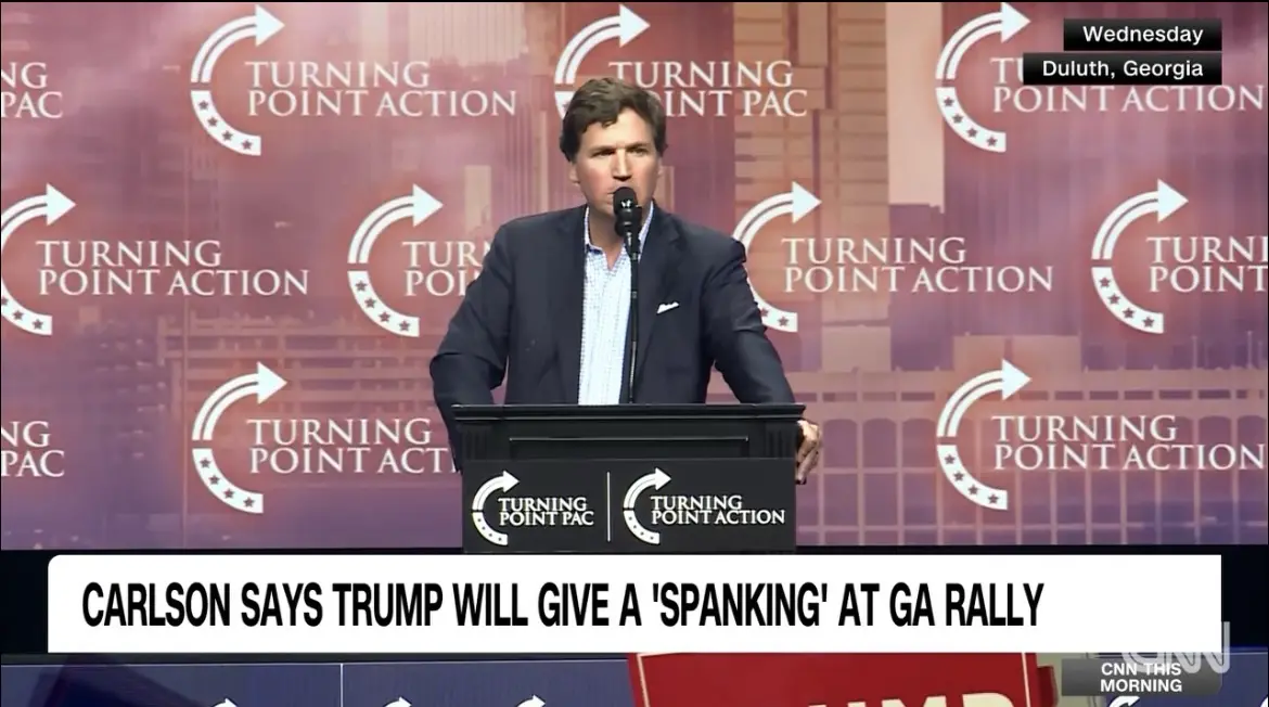 A man in a dark suit stands at a podium with 'Turning Point Action' logos on it, speaking into a microphone. Behind him is a…