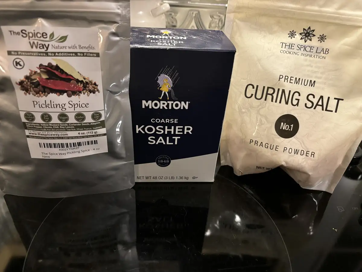Three kitchen salt products are arranged side-by-side on a black stovetop: a bag of 'The Spice Way' Pickling Spice, a box of…