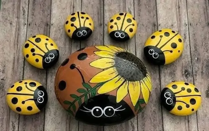 A large painted rock shaped like a sunflower with a ladybug face at the bottom sits surrounded by six smaller painted…