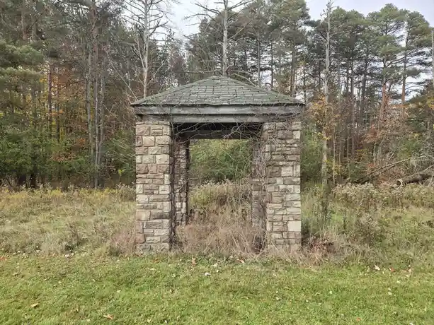 A stone gazebo with a wooden roof stands in an overgrown grassy field surrounded by autumnal trees. Bare vines and weeds…