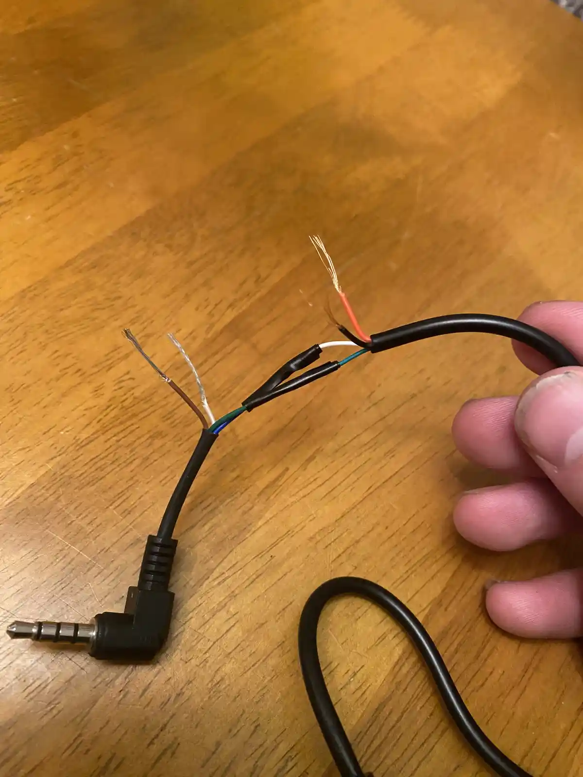 A hand holds a black audio cable with its wires stripped and exposed, showing a broken connection near the 3.5mm jack, which…