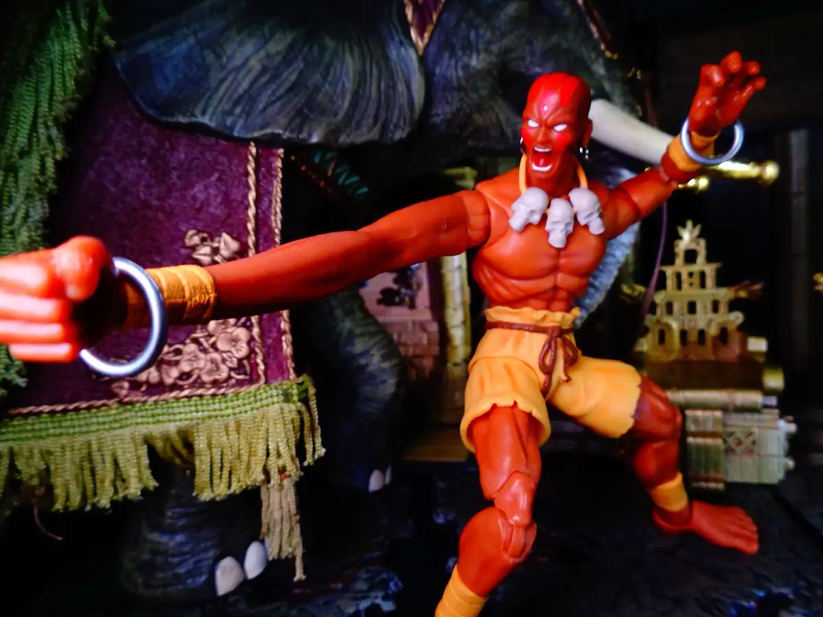 A red-skinned action figure with a muscular build, wearing orange shorts and skull necklaces, strikes a dynamic pose with…