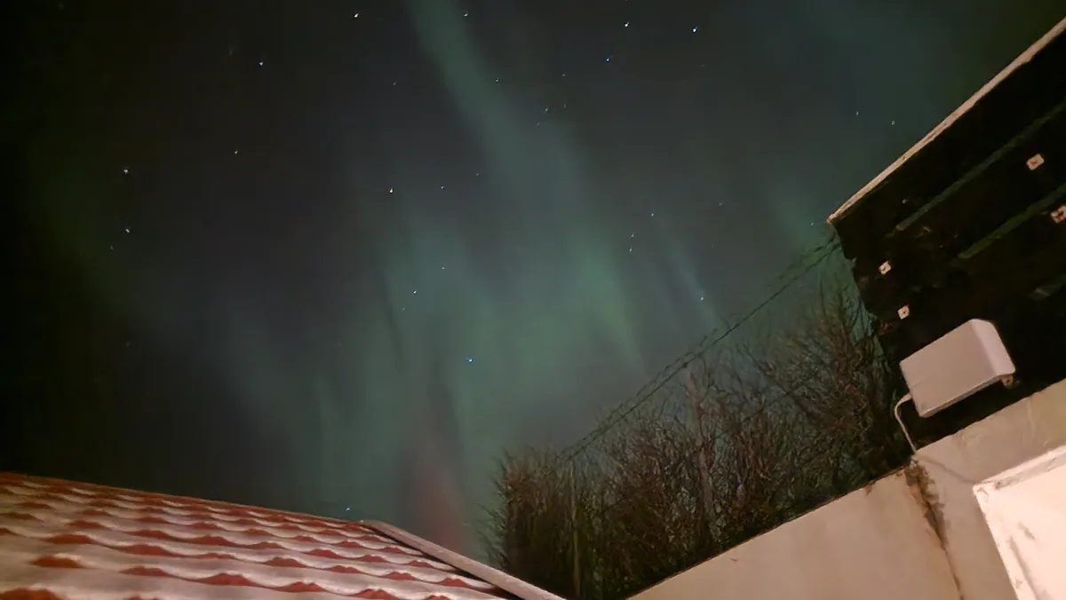 A low-angle nighttime photo shows a vibrant green aurora borealis dancing across a starry sky, framed by the roof of a…