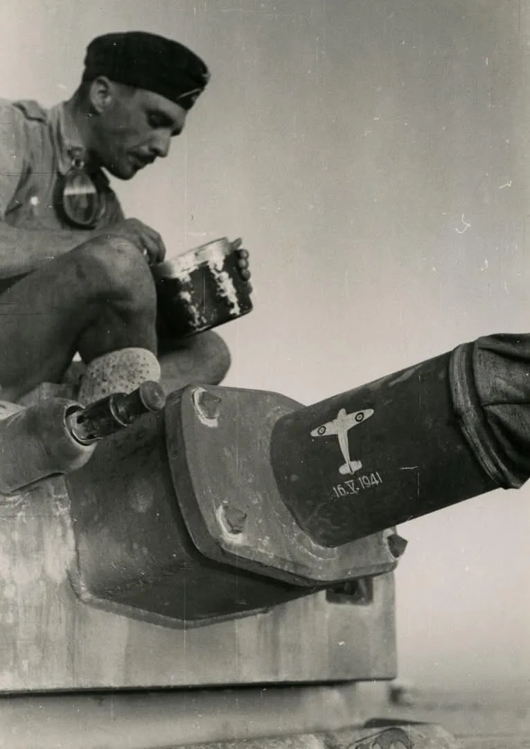 A black-and-white historical photo shows a soldier in military uniform sitting atop a tank, eating from a metal tin. The…
