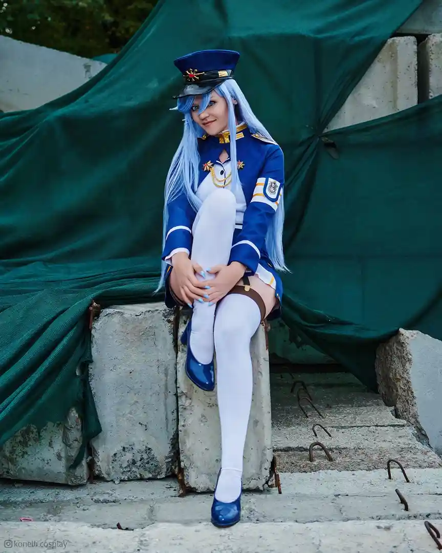 A cosplayer with long blue hair and a blue military-style uniform sits on a concrete block, smiling at the camera. The…