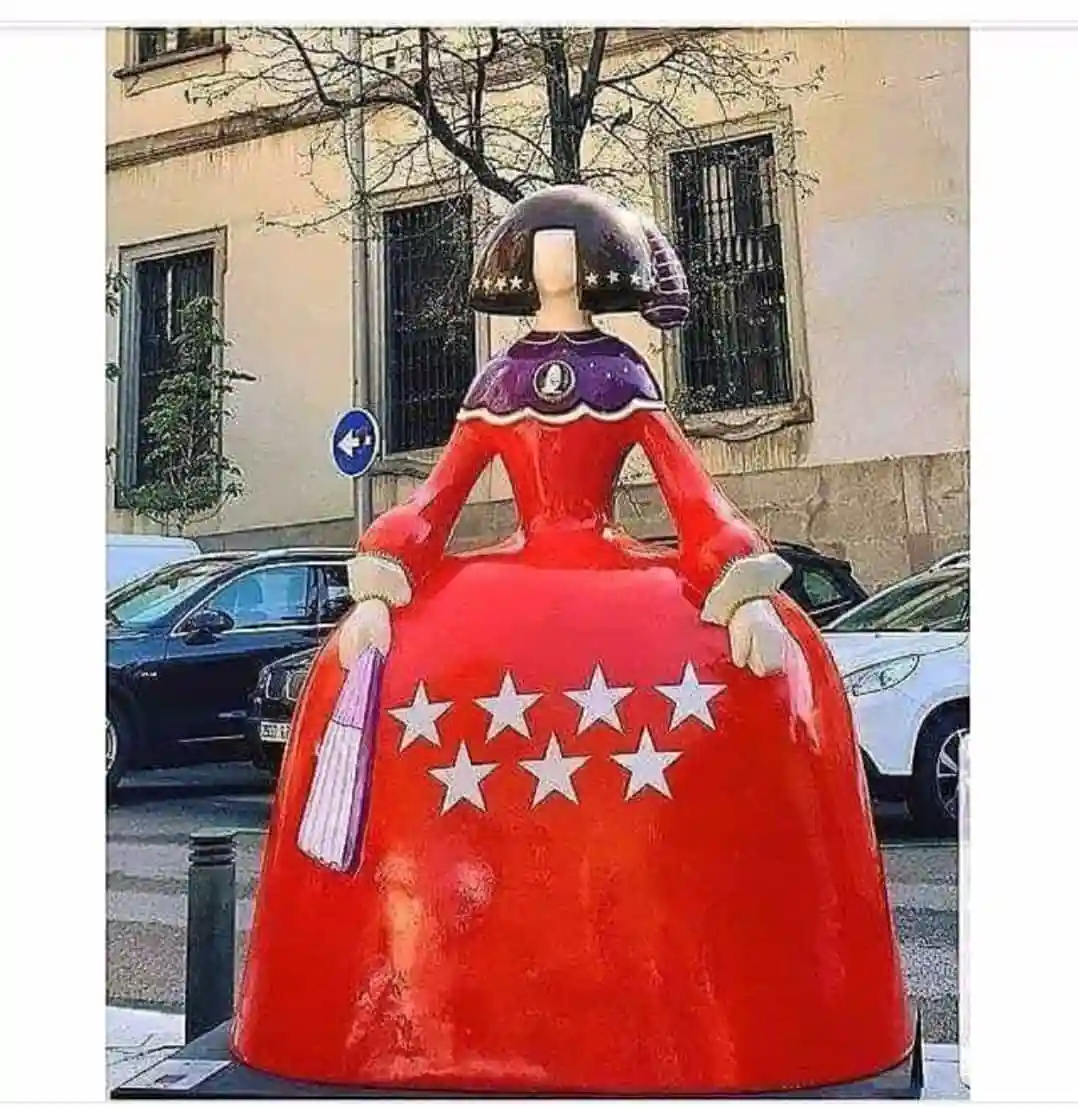 A large, stylized statue of a woman in a red dress with white stars and a purple collar stands on a city street, facing away…