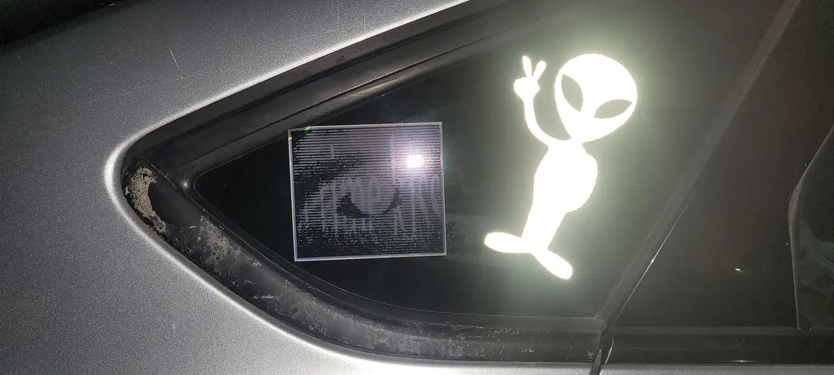 A close-up of a car's rear window shows a glowing white alien silhouette making a peace sign, next to a square sticker with…