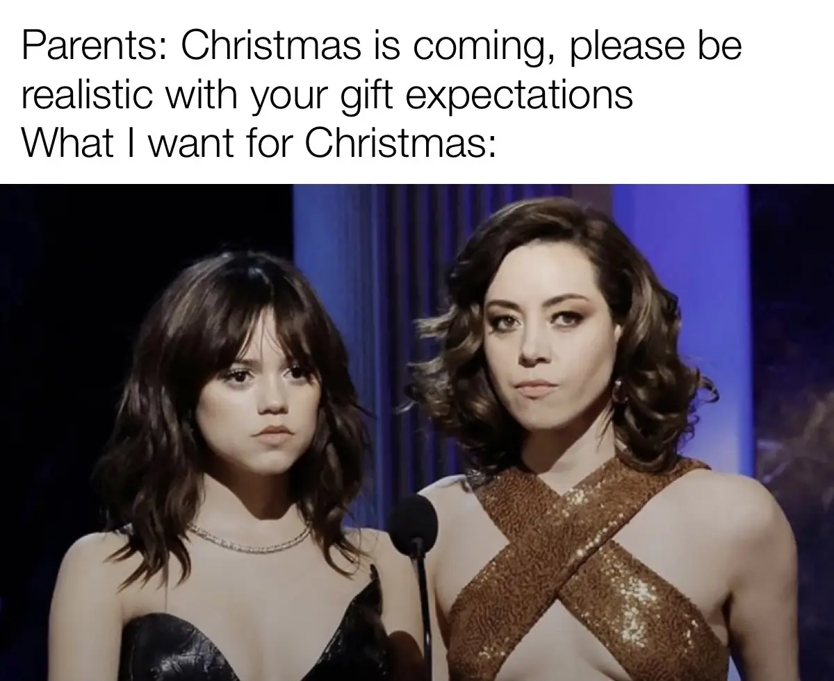 Two women stand side by side on a stage, both with serious, unimpressed expressions. The woman on the left wears a black…
