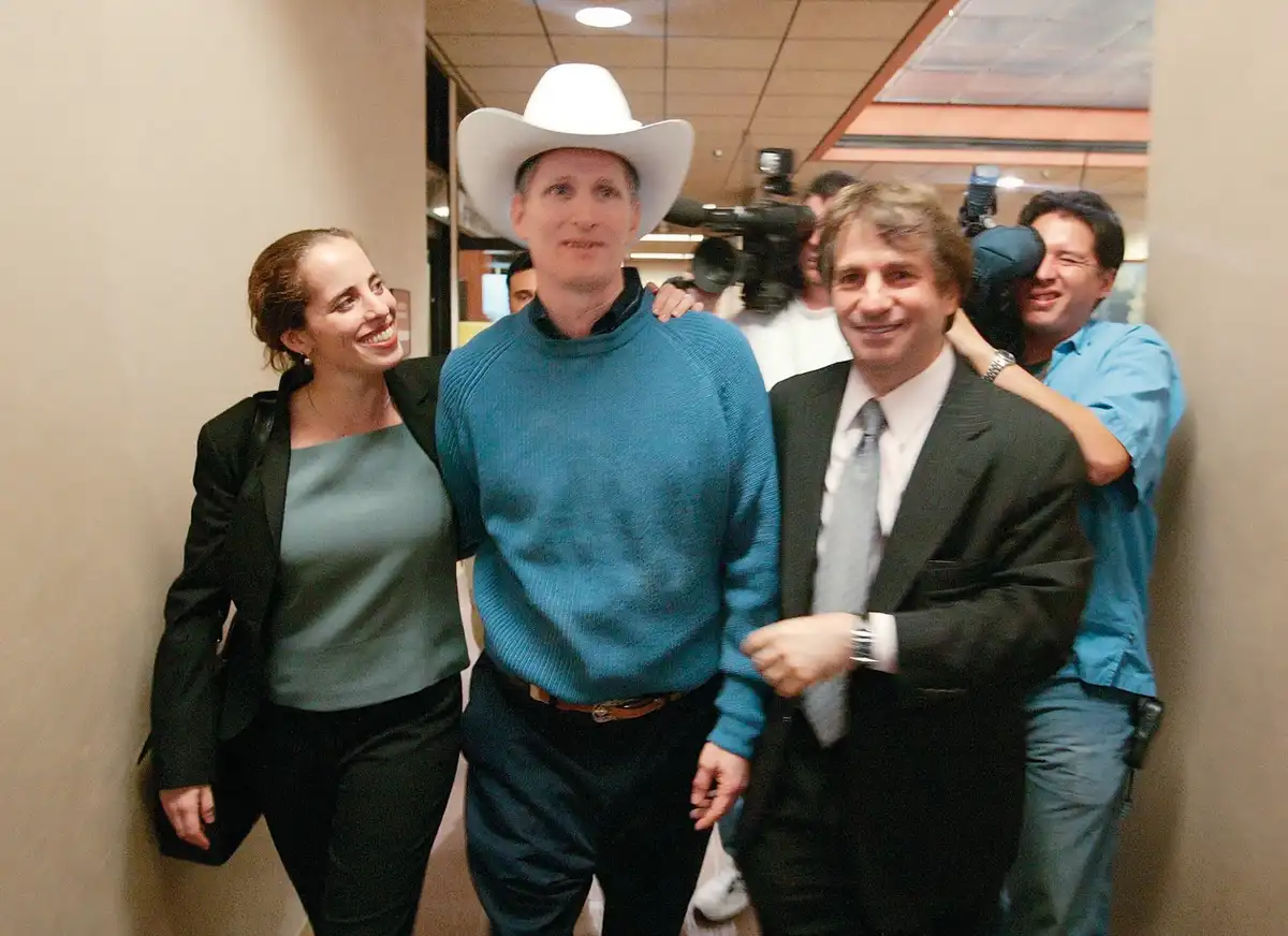 A man in a white cowboy hat and blue sweater stands in a hallway with two smiling people, one woman on his left and one man…