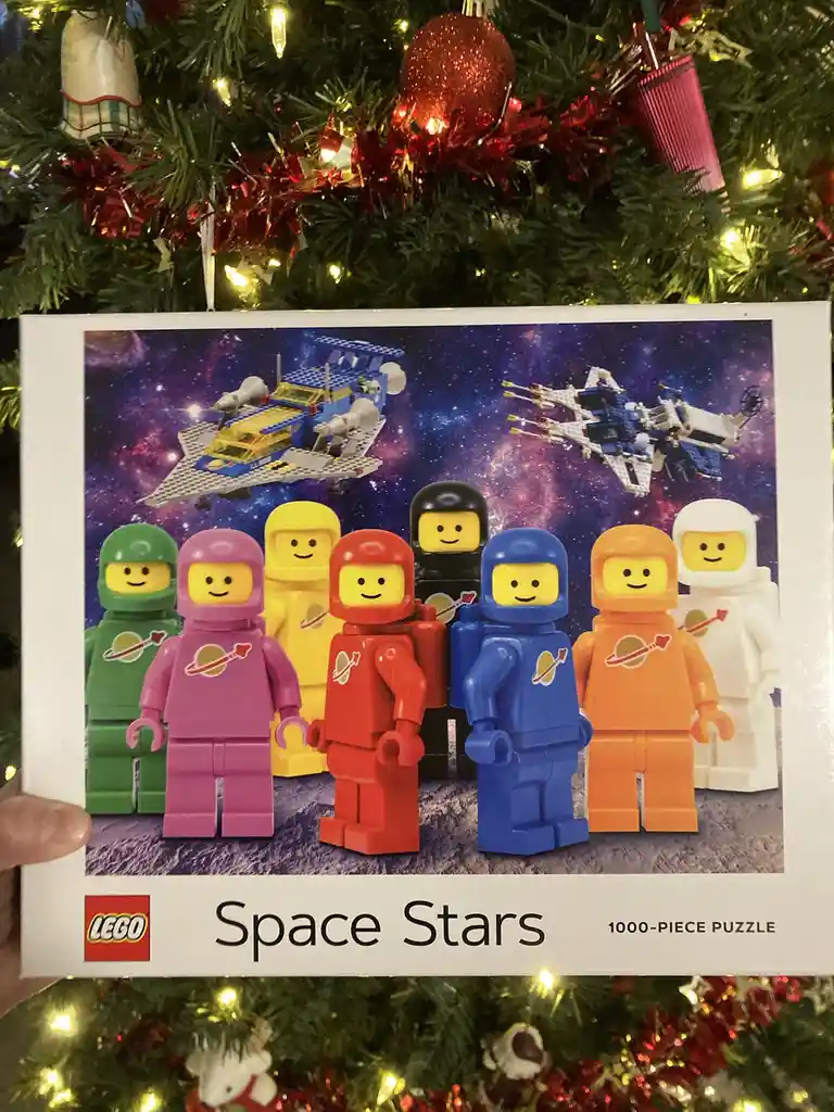 A hand holds a LEGO puzzle box titled 'Space Stars' featuring seven colorful astronaut minifigures standing on a rocky…