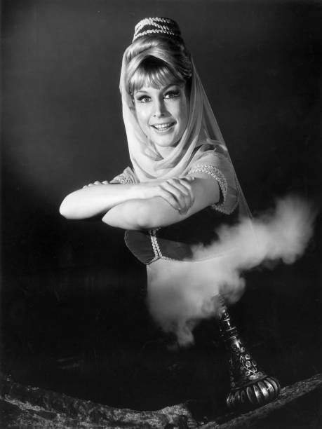 a black-and-white photo of a woman in a genie costume, smiling with arms crossed, standing beside a smoking magic lamp. The…