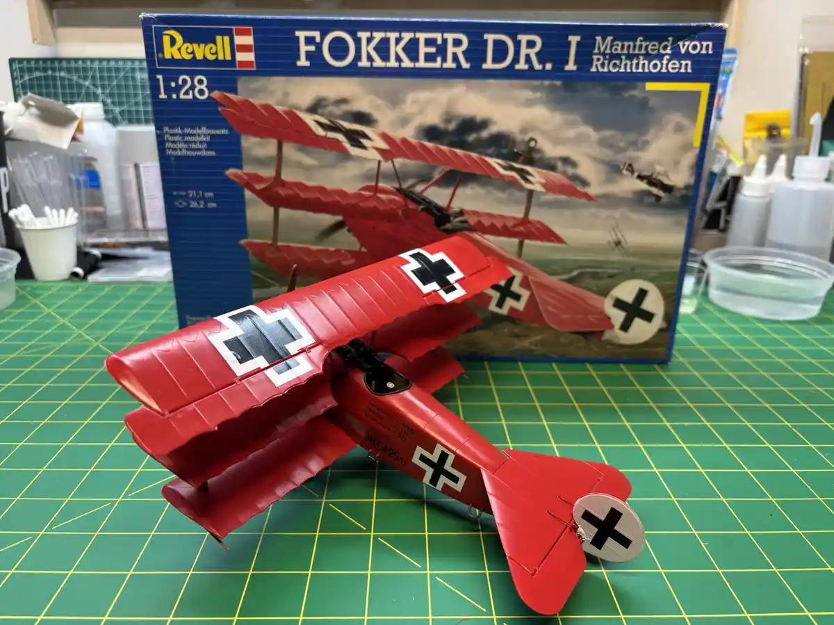 A completed red 1:28 scale plastic model of a Fokker Dr.I triplane, featuring black cross markings, sits on a green cutting…