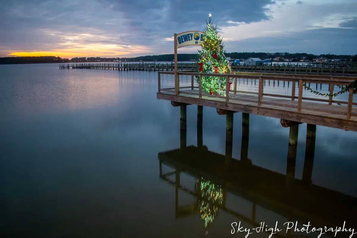 A festive Christmas tree adorned with lights and red bows stands on a wooden pier at dusk, with a sign reading 'DEWEY DE…