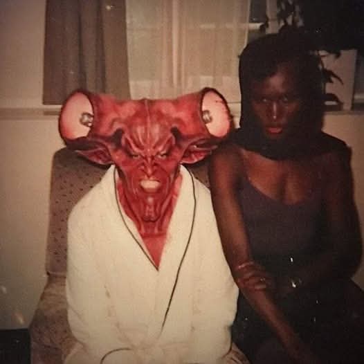 A person wearing a red, horned, demonic mask sits in a chair while another person sits beside them. The masked person wears…