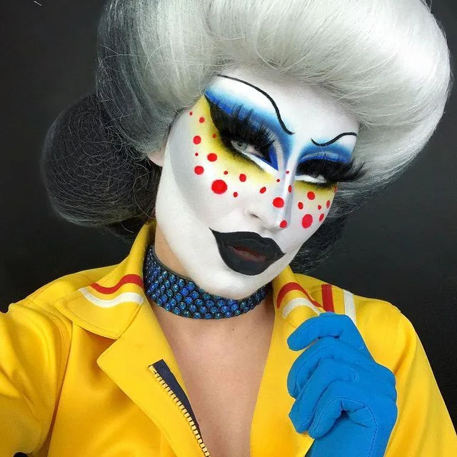 A person in drag with a white wig, bold blue and yellow face paint, red polka dots, black lipstick, and a yellow jacket…